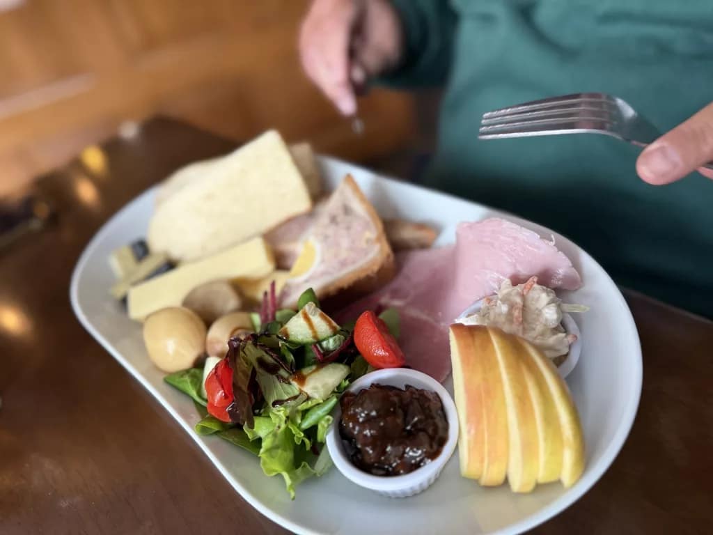 A traditional Ploughman’s lunch on a white plate with ham, cheddar cheese, pork pie with egg, bread, salad, apple slices, coleslaw, pickled onions, and chutney, ready to eat.