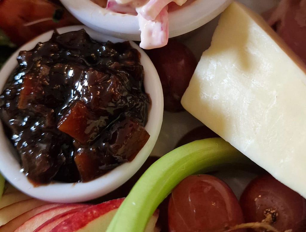 Close-up of a small white ramekin filled with chunky dark brown pickle beside a wedge of cheese, red grapes, celery, and apple slices on a Ploughman’s lunch plate.