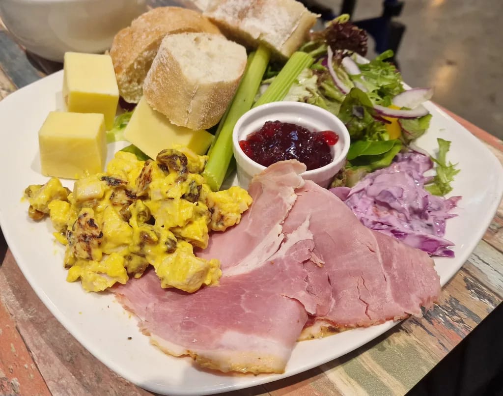 Ploughman’s-style lunch with sliced ham, coronation chicken, crusty bread, butter, celery, mixed salad, red cabbage slaw and a ramekin of chutney on a rustic table.