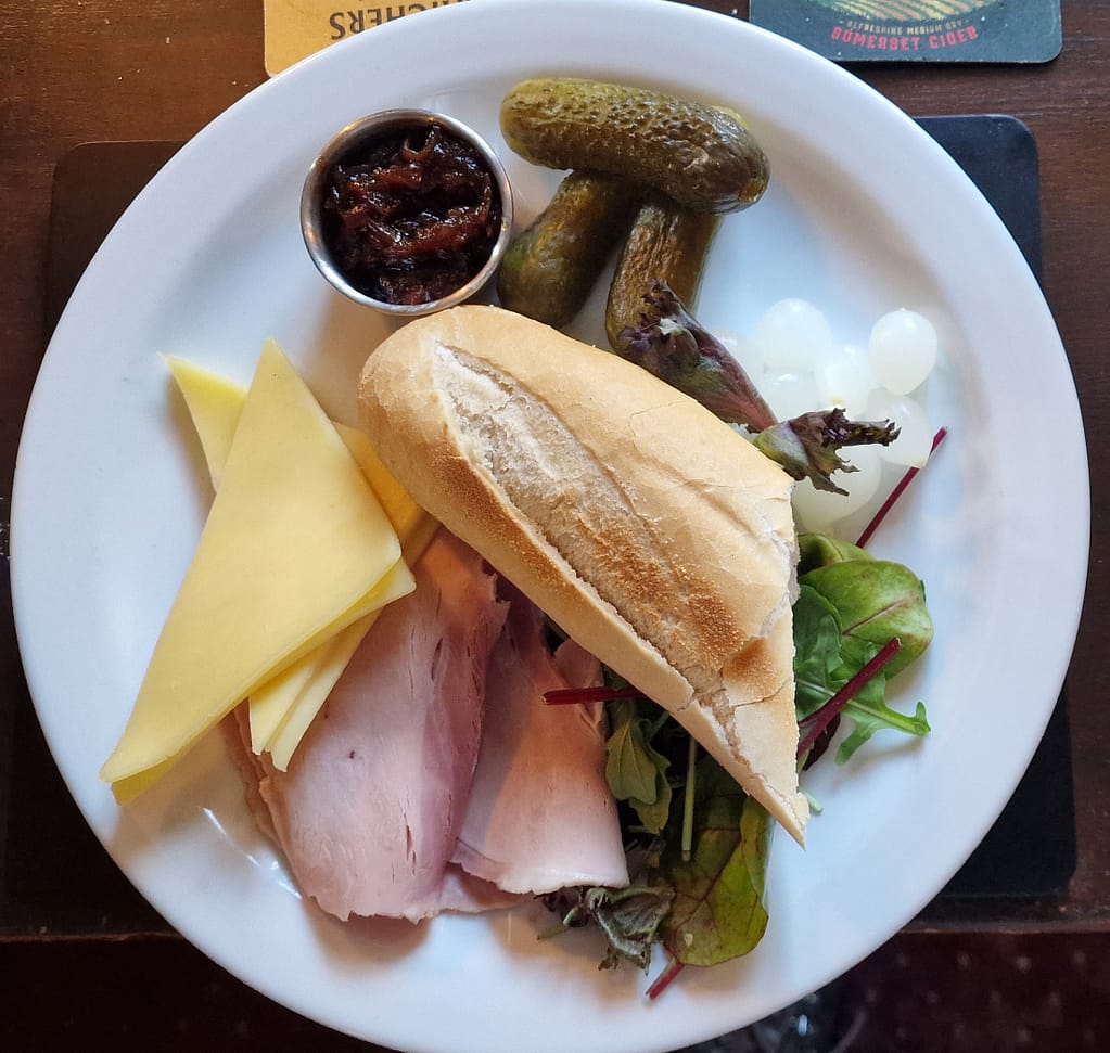 
The image is a top-down view of a classic Ploughman's lunch, served on a white plate. The meal includes a sliced, crusty baguette in the center, with two slices of ham and a few slices of mild cheddar cheese arranged neatly on the left side of the plate. On the right, there are three large pickled gherkins, a few pickled onions, and a small metal dish containing chutney. The dish sits on a base of fresh mixed greens, which adds a touch of color to the presentation. The plate is set on a dark wooden table, and the corner of a coaster is visible, hinting at the casual pub dining atmosphere. The clean, organized presentation emphasizes the simplicity and traditional elements of this British favorite.