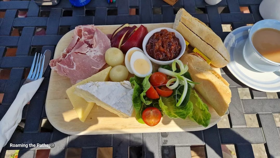 A traditional ploughman’s lunch served on a wooden board, featuring thick slices of ham, wedges of cheddar and brie cheese, a boiled egg, pickled onions, cherry tomatoes, lettuce, sliced red apple, and a generous portion of chutney. Two crusty baguette halves with butter packets sit beside the salad. A cup of tea in a white cup and saucer is placed on the black metal outdoor table next to the board. The setting is sunny and relaxed, suggesting an alfresco meal.
