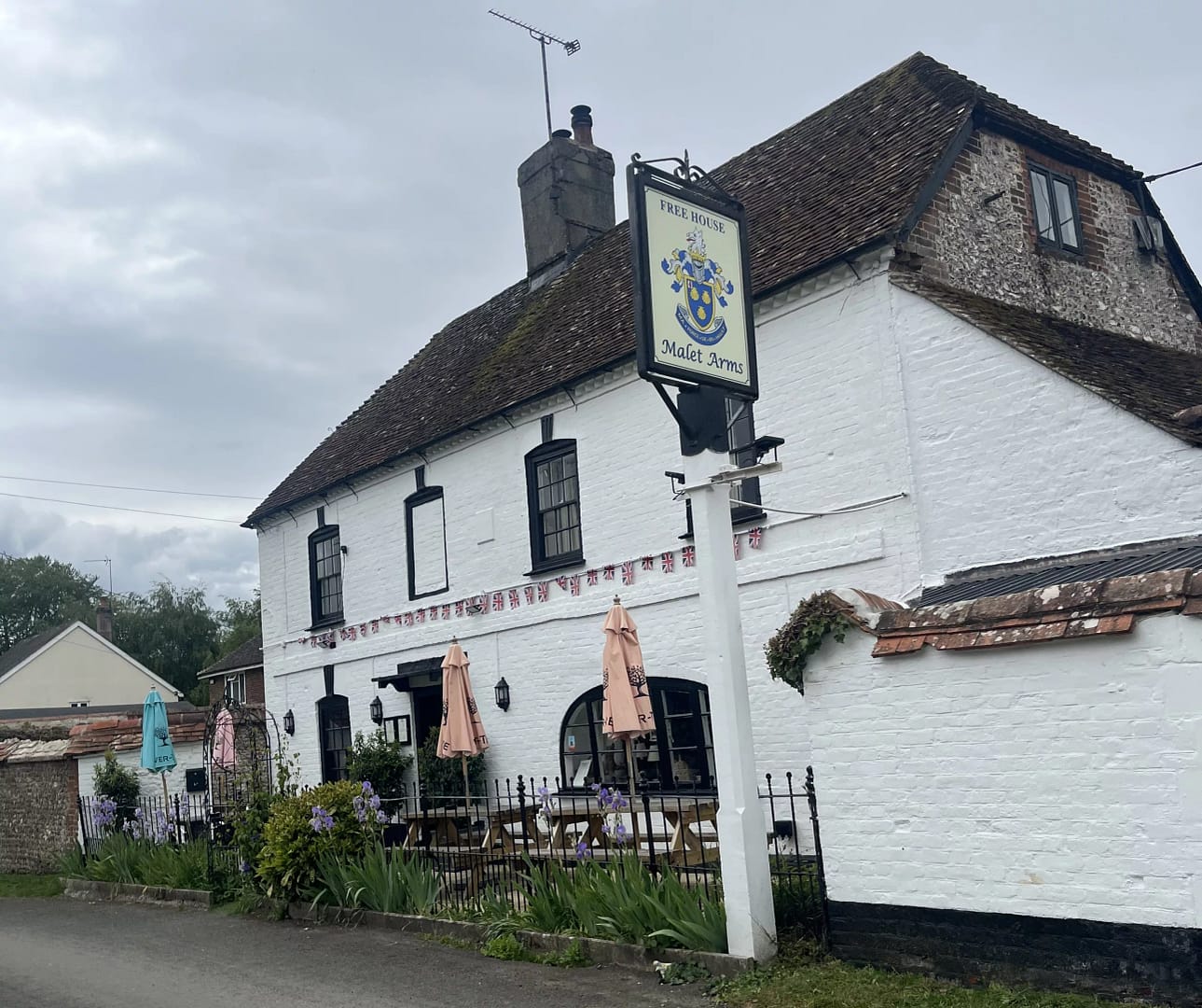 The exterior of the Malet Arms, a traditional white-painted country pub with black trim and a steep tiled roof. A hanging pub sign stands beside the building, with outdoor seating, umbrellas, flowers, and bunting decorating the frontage beneath a cloudy sky.