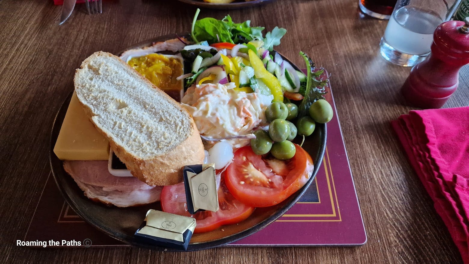 A plate of Ploughman's lunch featuring a slice of crusty bread, thick cheddar cheese, ham, sliced tomatoes, green olives, mixed salad greens, pickled onions, and coleslaw. The plate also includes a small portion of chutney and two wrapped butter portions. The meal is served on a dark plate set on a wooden table with a red napkin and condiments nearby.