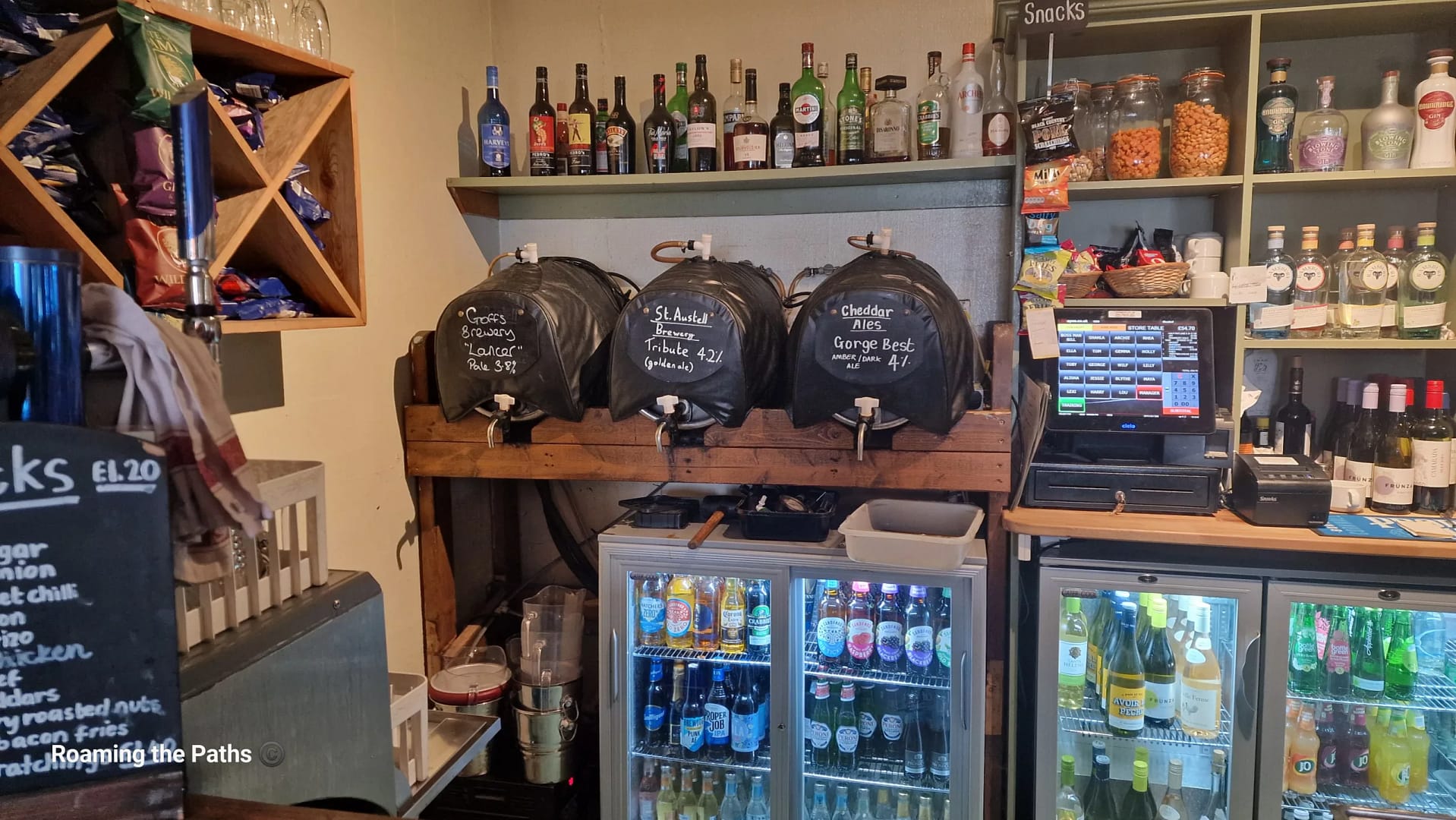 A traditional pub bar area featuring three casks of real ale labeled Groff’s Brewery Lancer, St Austell Tribute, and Cheddar Ales George Best. The bar is stocked with bottled drinks, spirits, snacks, and a touchscreen till, with shelves displaying crisps, wine, and local gin