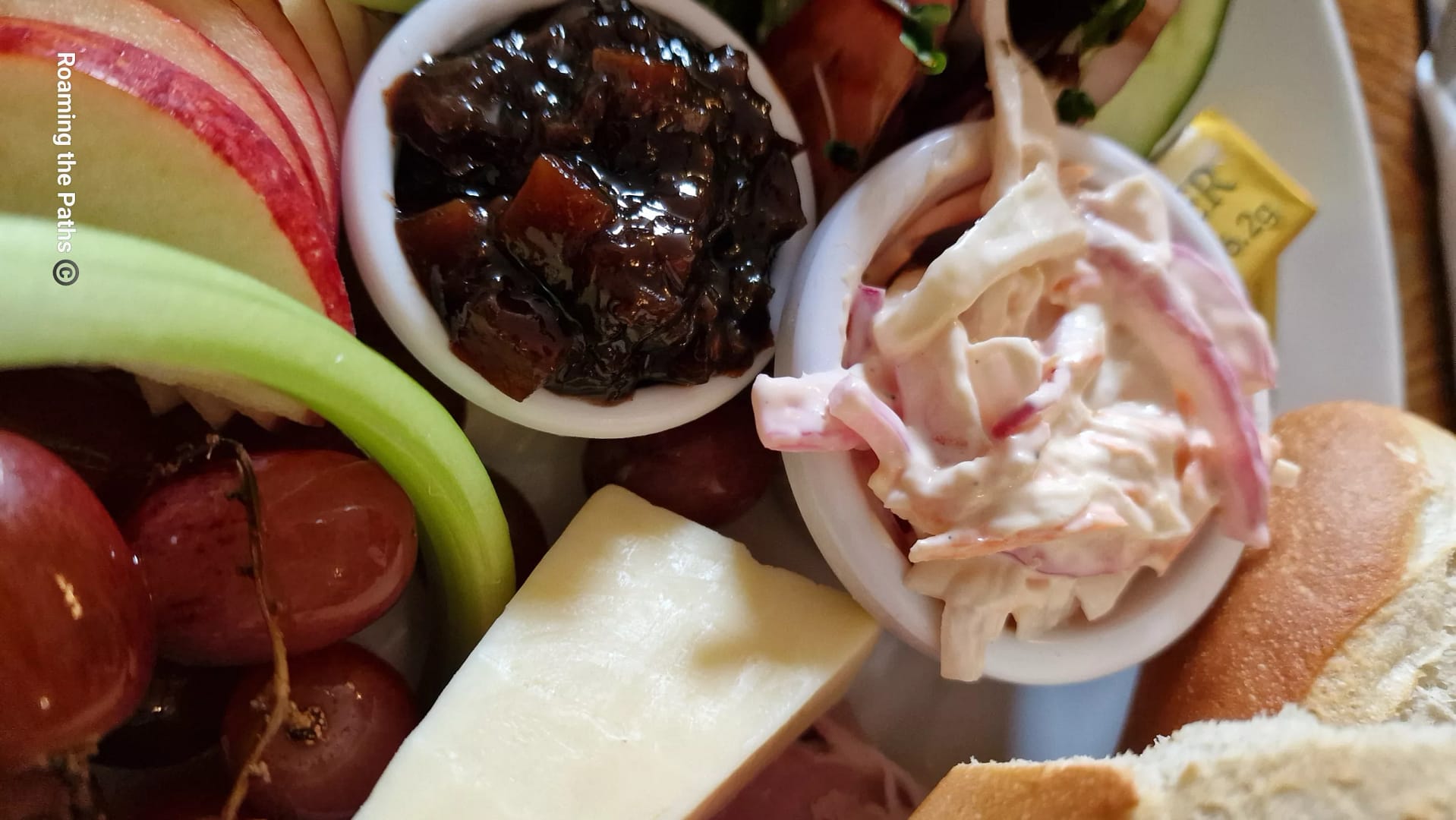 Close-up of a Ploughman’s lunch showing small pots of creamy coleslaw and chunky dark pickle, surrounded by cheese, sliced apple, celery, grapes, and crusty bread.