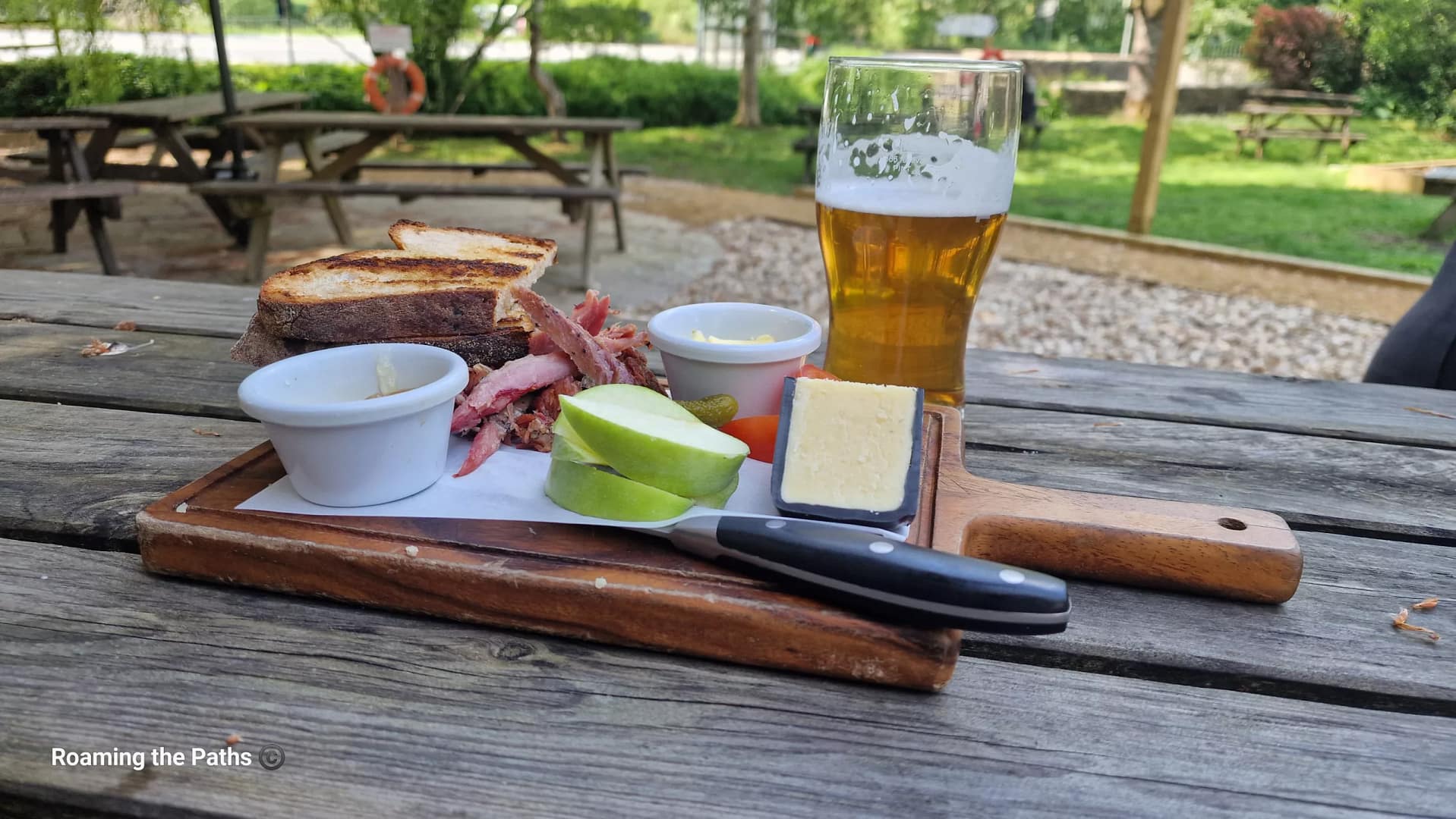 A rustic Ploughman’s lunch served on a wooden board at an outdoor pub garden beside a canal. The board includes grilled bread, thick-cut ham, a wedge of cheese, apple slices, pickles, butter, and salad, accompanied by a pint of golden beer. Wooden picnic benches and leafy greenery create a relaxed summer setting in the background.