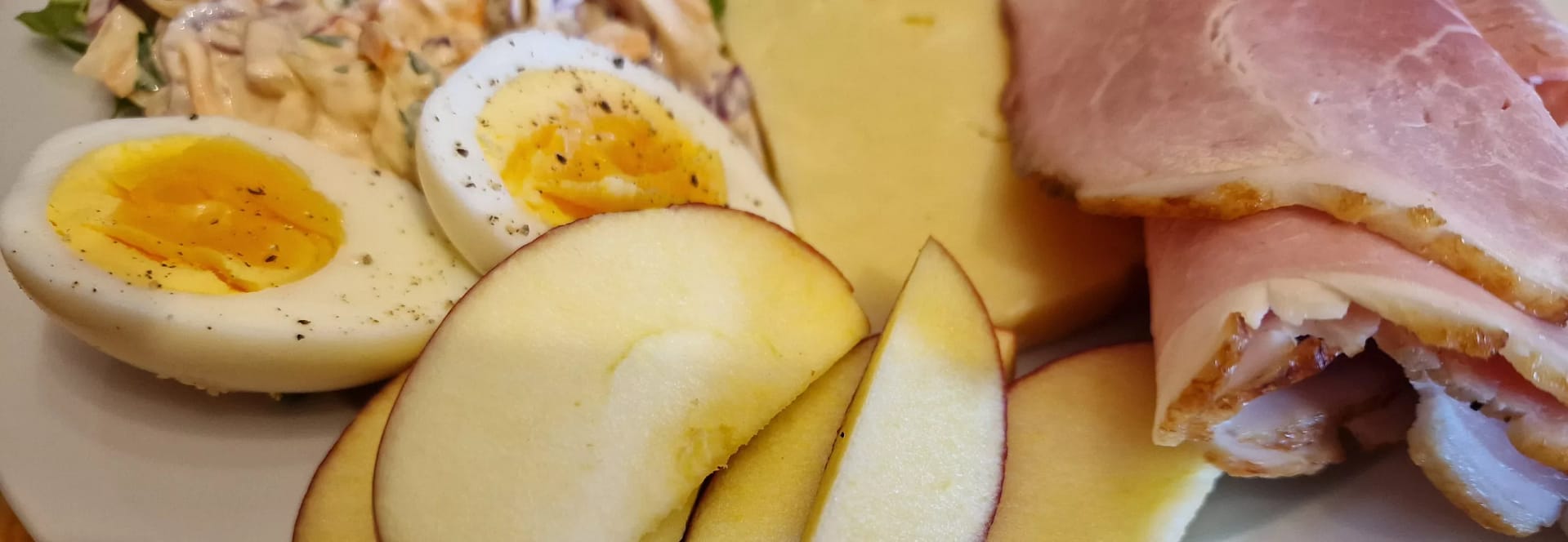 Close-up of a Ploughman’s lunch showing two halves of a hard-boiled egg with pepper, slices of red apple, folded slices of ham with a browned edge, and a wedge of cheddar cheese, with creamy coleslaw just visible in the background.