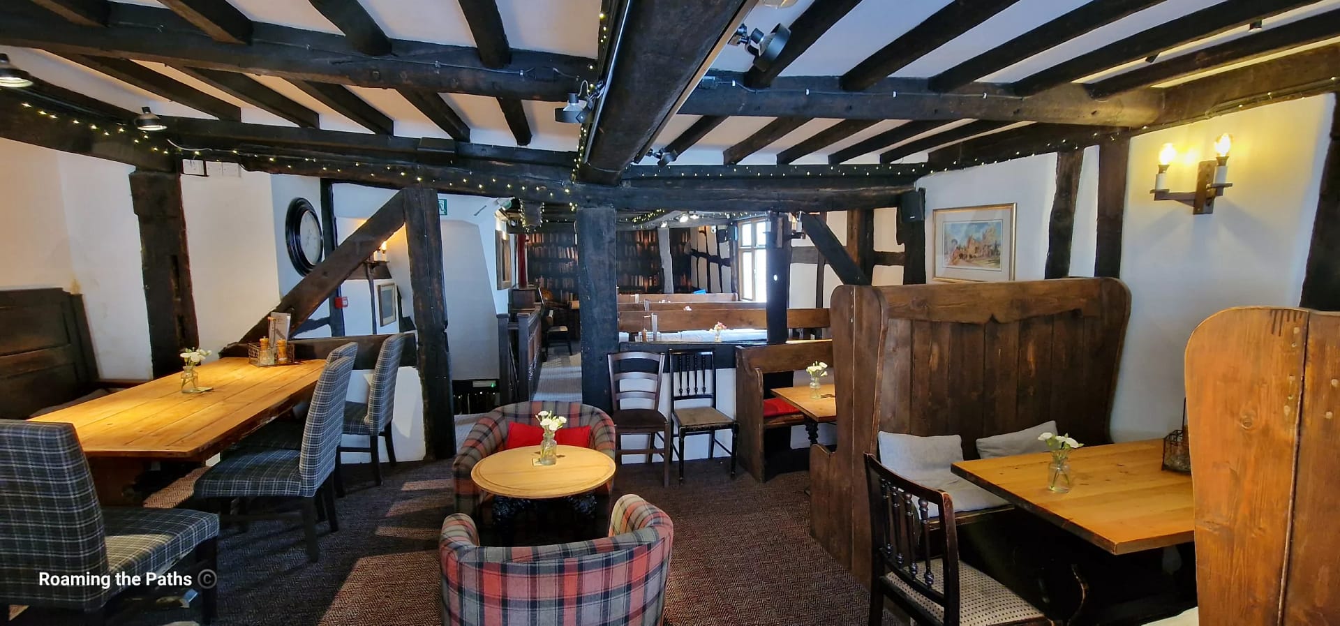 A cosy dining area inside a historic timber-framed pub. Exposed black oak beams, wooden booths, tartan armchairs, and polished wooden tables create an inviting atmosphere, with small flower vases and soft lighting adding to the traditional character.