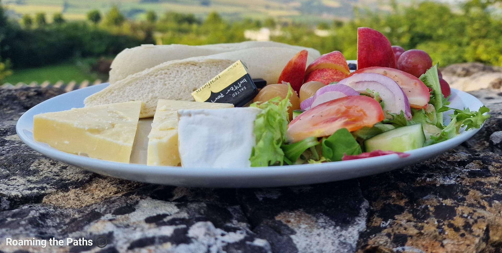 Ploughman's lunch served on a plate with cheese, bread, salad, and fruit, placed on a stone wall with a scenic countryside view in the background