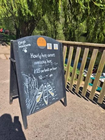 Chalkboard sign advertising canoe hire by a riverside pub, with “dogs welcome” message and wooden railing in sunshine.