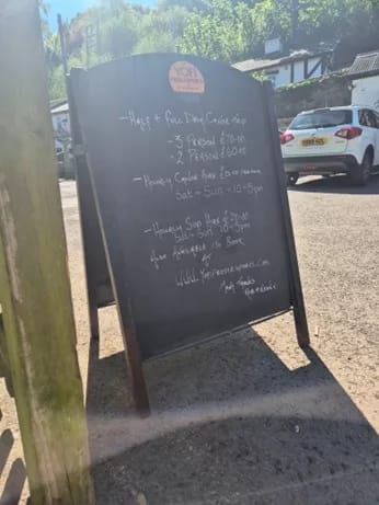 Chalkboard sign outside a pub listing canoe hire prices and opening times in a sunny car park.