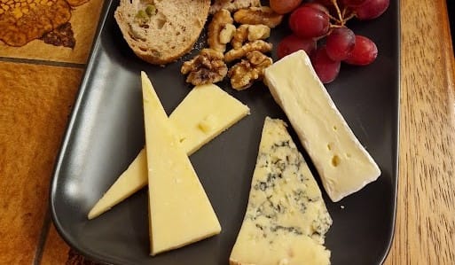 This image displays a neatly organized cheese platter on a rectangular black plate. The selection includes three cheeses: a firm cheddar cut into long triangles, a creamy wedge of brie, and a piece of blue cheese with distinct veining. Alongside the cheeses, there are slices of artisan bread, a handful of walnuts, and a small bunch of red grapes. The table beneath has a warm wooden texture, contributing to a rustic, inviting presentation. The arrangement emphasizes simplicity while offering a classic combination of flavors and textures for a cheese board.