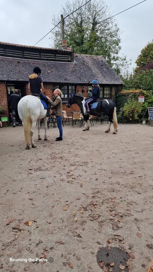 This image captures a charming outdoor scene in front of a rustic brick building, likely part of a countryside café or farm shop. Two riders on horseback are stopped outside, with one woman interacting with the riders, possibly adjusting tack or giving instructions.
The horses, one white and the other black and white, stand calmly as they wait, and both riders are wearing helmets and casual riding attire. The ground is scattered with dry leaves, adding an autumnal touch. The building in the background has a tiled roof, ivy-covered walls, and an inviting setup with tables and chairs, suggesting it’s a popular stop for both locals and passersby, including those on horseback. This peaceful scene reflects the leisurely pace and close-to-nature lifestyle typical of rural settings.