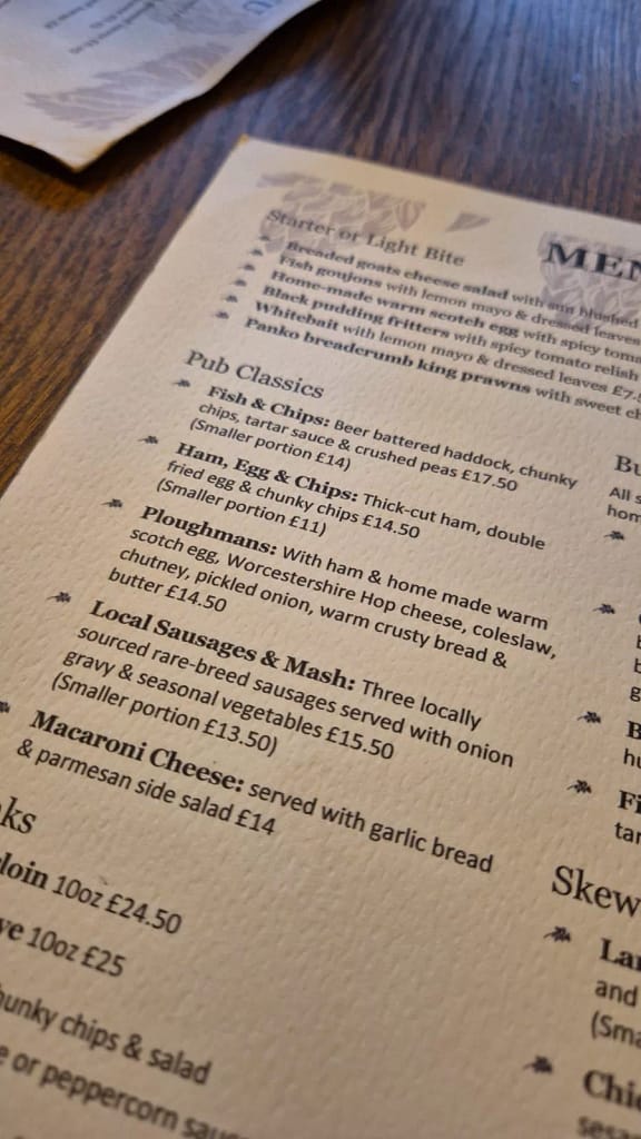 Close-up of a printed pub menu from The Alma Inn, resting on a wooden table. The visible section features the "Pub Classics" and "Starter or Light Bite" categories. Dishes include beer-battered fish and chips, ham with eggs and chunky chips, a Ploughman's lunch with ham, warm Scotch egg, Worcestershire Hop cheese, coleslaw, chutney, pickled onion, crusty bread and butter, local sausages and mash with onion gravy, and macaroni cheese with garlic bread and salad. Prices range from £11 to £17.50, with smaller portion options listed. The layout is clean with classic serif fonts on cream textured paper.