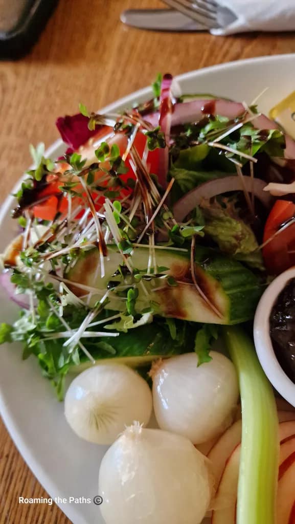 Close-up of a fresh salad with mixed greens, cucumber, tomato, red onion, and sprouts drizzled with dressing, beside pickled onions and celery on a Ploughman’s lunch plate.