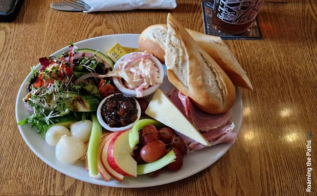 A traditional Ploughman’s lunch served on a white plate, featuring crusty baguette slices, thick-cut ham, cheddar cheese, pickled onions, apple slices, grapes, celery, chutney, coleslaw, and a fresh salad with mixed leaves and sprouts. A pint of cider sits on the wooden pub table beside the plate.