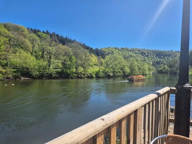 River view from a pub terrace with a boat passing and wooded hills in bright sunshine.