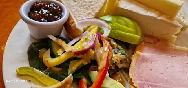 This image shows a close-up of a Ploughman’s lunch served on a white plate. The plate features a variety of classic components, including thick slices of cheddar and brie cheese, and a portion of ham with a visible pink, succulent texture. A small dish of dark, chunky chutney is positioned in the upper left corner of the plate. Fresh slices of green apple are arranged alongside the cheeses, adding a crisp, sweet element.
In the center, there is a vibrant mixed salad with colorful strips of red and yellow bell peppers, cucumber, spinach leaves, and thinly sliced red onions, all drizzled with a mustard dressing. The salad appears fresh and lightly coated, adding a tangy contrast to the other elements. There is also a portion of fresh, thick-sliced white bread or baguette, slightly visible under the salad and cheese. The wooden table beneath the plate adds to the rustic, comforting feel of this traditional British pub meal.