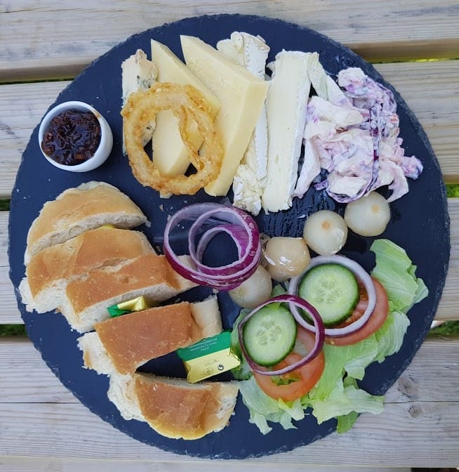 A circular slate platter holds a traditional Ploughman’s lunch arranged beautifully. On the left, several thick slices of fresh bread are served with butter pats. In the center, a small mound of pickled onions, rings of red onion, and a fresh salad of lettuce, cucumber, and tomato add color. The cheese selection includes cheddar, brie, and a wedge of another hard cheese, topped with a crispy onion ring. To the right, there's a creamy coleslaw and a small pot of chutney. The presentation gives a rustic feel, set against a wooden picnic table backdrop.