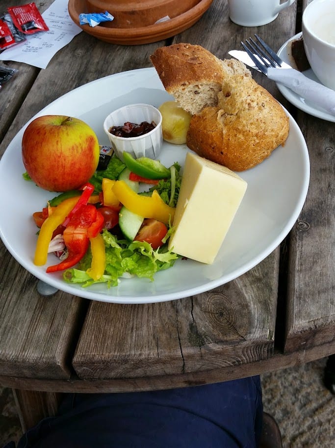 A large white plate features a classic Ploughman’s lunch. It includes a wedge of cheddar cheese, a crusty whole grain bread roll, and a fresh apple. A colorful salad of lettuce, tomato, cucumber, and bell peppers is on the side, along with a small cup of chutney, a pickled onion, and butter. The dish is bright and vibrant, with a balance of fresh produce and hearty components.