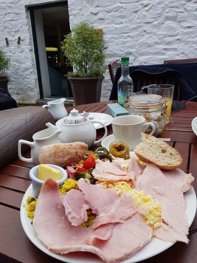 A Ploughman’s lunch served outdoors at Colby Woodland Garden, featuring an inviting mix of sliced ham, grated cheese, and a fresh salad with vibrant tomatoes, cucumbers, and peppers. The plate includes a small jar of pickles, a crusty bread roll with a pat of butter, and a side of couscous. The lunch is accompanied by a cup of tea, complete with a colorful teapot, a milk jug, and a sugar jar with cubes. The outdoor seating captures the peaceful ambiance, with greenery and white-washed walls of a garden café in the background, giving a charming, rustic feel.