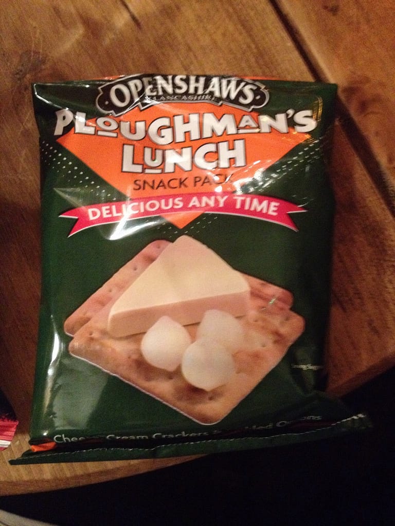 A close-up image of a snack pack labeled "Openshaws Ploughman's Lunch," featuring a green, orange, and black design. The slogan "Delicious Any Time" is prominently displayed on a red banner below the product name. The packaging suggests a convenient, ready-to-eat version of the traditional Ploughman's lunch, offering an easy snack option.