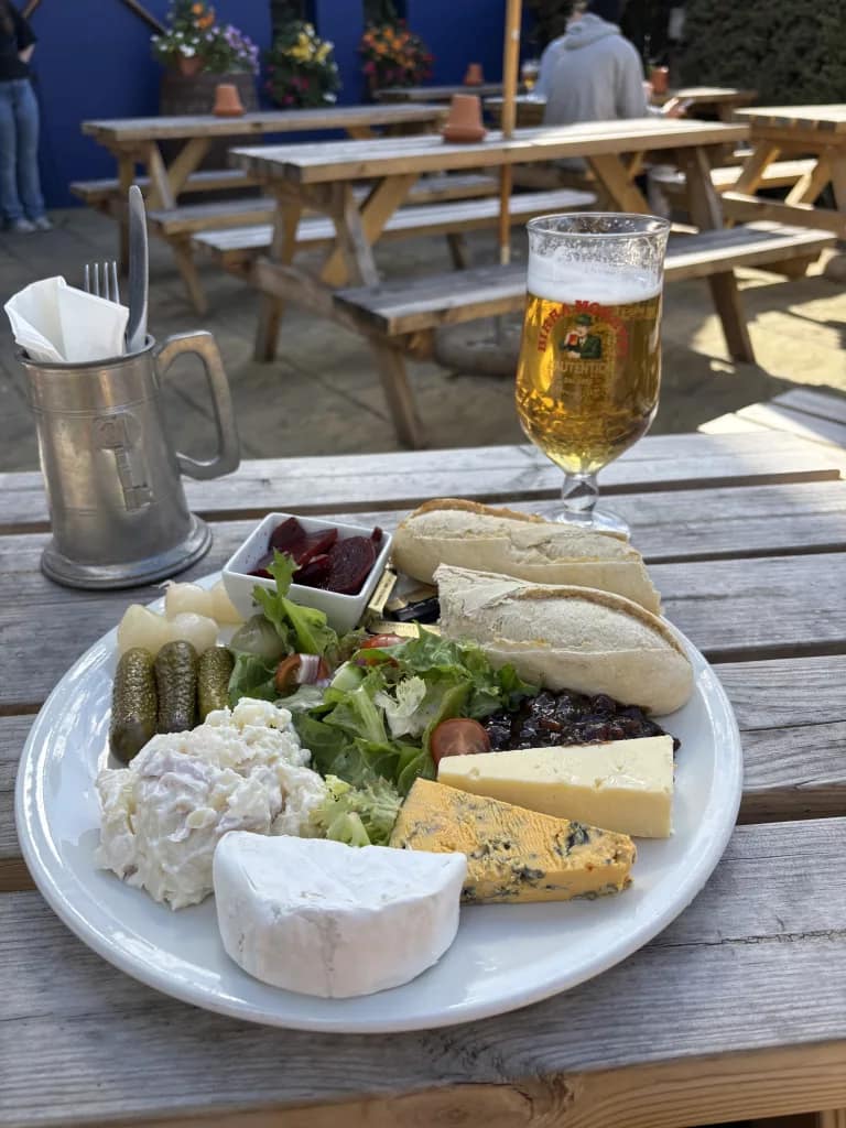 Ploughman’s lunch served outdoors on a wooden table: plate with cheeses, salad, pickles, potato salad, bread, chutney and beetroot, with a glass of lager beside it.