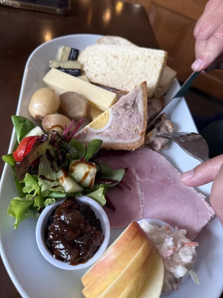 A traditional Ploughman’s lunch served on a white plate, featuring ham, pork pie with egg, cheddar cheese, salad, pickled onions, apple slices, coleslaw, bread, and a small pot of pickle.