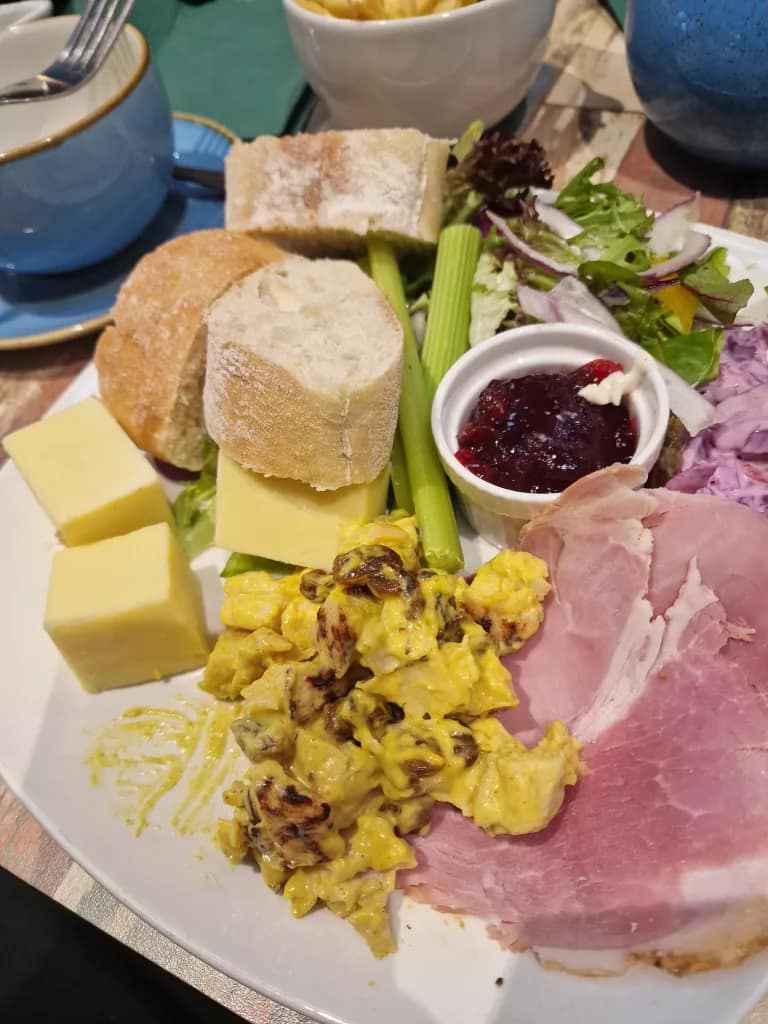 Ploughman’s-style lunch with sliced ham, coronation chicken, crusty bread, cheese, celery, mixed salad, red cabbage slaw and a ramekin of chutney on a café table.