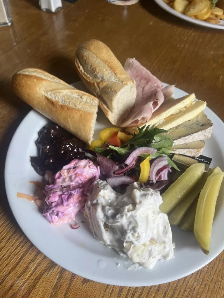 A pub-style Ploughman’s lunch served on a white plate on a wooden table. The meal includes crusty baguette, slices of ham, several cheeses including blue cheese, pickled gherkins, mixed salad leaves with red onion and peppers, creamy potato salad, pink coleslaw, and Branston Pickle.