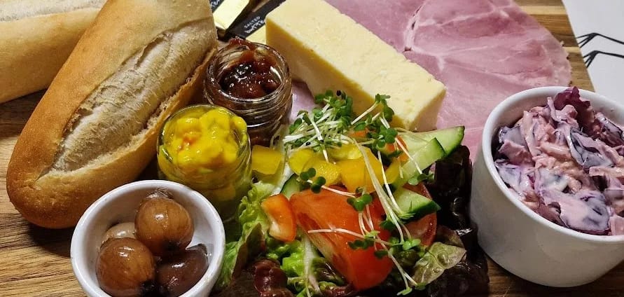 The image features a beautifully presented Ploughman’s Buffet spread, showcasing a variety of fresh, locally-sourced ingredients. Central to the platter is a thick wedge of rich, creamy cheddar cheese, accompanied by a couple of slices of succulent, pink-cured ham. There’s a golden-brown baguette, halved and crusty, invitingly ready to be filled or enjoyed alongside the accompaniments.

Surrounding the main components are small jars and bowls filled with traditional Ploughman’s condiments and sides. A jar of tangy, bright yellow piccalilli sits next to a glossy jar of rich, dark chutney. A separate bowl contains crisp pickled onions, their golden-brown hue glistening. Another bowl houses a vibrant coleslaw with a creamy, pinkish dressing, enhanced by beetroot.

Adding a splash of color and freshness to the plate is a neatly arranged salad, with a mix of leafy greens, cucumber slices, juicy red tomatoes, and vibrant yellow peppers. It’s topped with a scattering of fresh sprouts for an extra touch of texture and color.

The arrangement sits on a wooden board, lending a rustic and inviting feel, and perfectly represents the essence of a traditional yet elevated Ploughman’s Buffet.