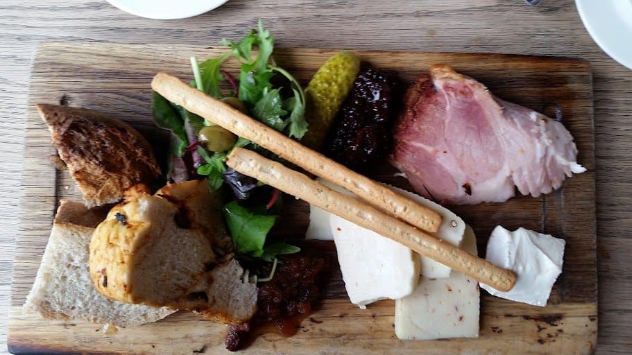 A wooden board is laid with a generous Ploughman's lunch featuring thick slices of ham, assorted cheeses including soft white cheese and a peppery variety, crusty bread, breadsticks, chutney, a pickled gherkin, and a fresh salad with leafy greens and olives. The rich textures of the bread and the smooth cheeses contrast beautifully with the sharp chutney and briny olives. The presentation is rustic, offering a hearty, delicious meal perfect for a café setting.