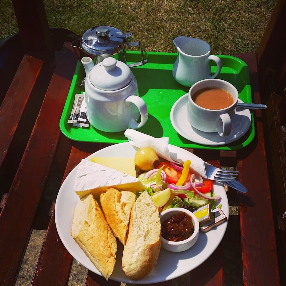 A vibrant outdoor setting featuring a traditional Ploughman’s lunch on a white plate. The meal includes two large slices of fresh baguette, a wedge of Brie, a slice of Cheddar, a small pot of chutney, a pickled onion, and a colorful salad of mixed greens, peppers, onions, cucumber, and tomato. A napkin-wrapped fork and knife rest beside the salad. In the background, a green tray holds a white teapot, a cup of tea with milk, a small milk jug, and sugar packets, highlighting a classic afternoon tea experience. The wooden table is bathed in sunlight, suggesting a pleasant, leisurely meal outdoors.