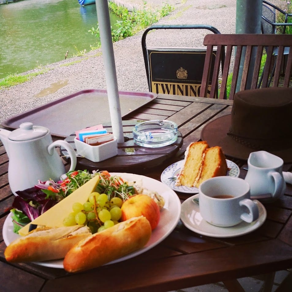The image shows a delightful Ploughman’s lunch served at an outdoor café table by the water. The meal includes: A wedge of cheddar cheese. A small baguette, halved. Fresh green grapes and an apple. A mixed salad with greens and shredded vegetables. A small serving of coleslaw. Beside the plate are a pot of tea, a teacup with milk jug, a slice of sponge cake on a separate plate, and a hat resting on the table. The peaceful riverside setting creates a serene and cozy atmosphere, perfect for enjoying a relaxed lunch.