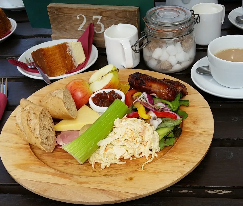 A wooden platter holds a classic Ploughman’s lunch with a variety of components. In the foreground, there is a crisp, fresh celery stick and a serving of creamy coleslaw. A few slices of rustic brown bread are placed alongside generous wedges of cheddar cheese and ham. To the left, sliced apple adds a fresh touch, and next to it, there’s a small bowl of dark chutney. The salad includes mixed greens, thinly sliced red onions, yellow and red bell peppers, and cucumber. On top of the salad sits a grilled sausage. The background features a cup of tea, a jar of sugar cubes, and a slice of sponge cake with a layer of frosting, all sitting on a dark wooden table. A rustic wooden table number "23" is displayed, indicating the table setting in an outdoor or café environment.