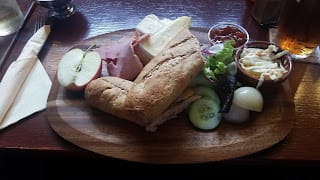 A wooden serving board holds a traditional Ploughman's lunch featuring a whole wheat baguette, slices of ham, cheddar cheese, a half apple, pickled onions, cucumber slices, mixed salad greens, and a small dish of coleslaw. A portion of chutney is placed alongside, completing the hearty meal. A pint of beer is visible in the background, set on a wooden table at The Lamb Inn, Axbridge.