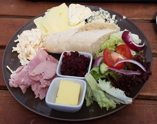 This image shows a plate featuring a classic ploughman’s lunch. The meal consists of several traditional elements including: A portion of sliced ham. A slice of crusty white bread or baguette. A wedge of butter. Slices of cheese (one appears to be cheddar, and another possibly a soft cheese or brie). A small portion of coleslaw. Some chutney or relish in a small dish. A fresh side salad containing lettuce, tomato, cucumber, and red onion. This type of meal is typically served as a hearty cold lunch, often with a variety of cheeses, meats, and pickles.
