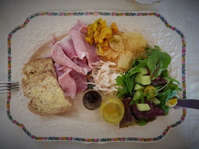 This image shows a traditional Ploughman’s lunch served on a rectangular floral-patterned plate. The meal includes slices of thick-cut pink ham, coleslaw with visible shredded vegetables, and a small mound of crispy potato chips or crisps. Accompanying these is a mixed salad of fresh greens, cucumber, and beetroot, with a drizzle of salad dressing placed in a small glass container. There is also a serving of bright yellow pickles, likely piccalilli, and a dark chutney in a tiny glass jar. The plate features two slices of buttered wholegrain bread. The arrangement is tidy, with distinct sections for each component, and the colorful ingredients stand out against the white plate, evoking a rustic yet appealing meal presentation