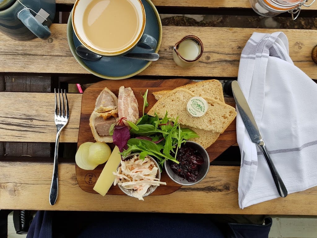 A flatlay of a Ploughman’s lunch served on a wooden board at The Swan Inn, Rowberrow. The meal includes slices of pork pie with a golden, crispy crust, a block of cheddar cheese, a pickled onion, a small portion of coleslaw, mixed salad leaves, and a dollop of chutney. The board also features two slices of brown bread with a small dish of butter. On the table, there's a blue cup of tea with milk in a matching saucer, a small jug of extra milk, a knife and fork, and a neatly folded white napkin. The rustic setting is enhanced by the wooden picnic-style table.
