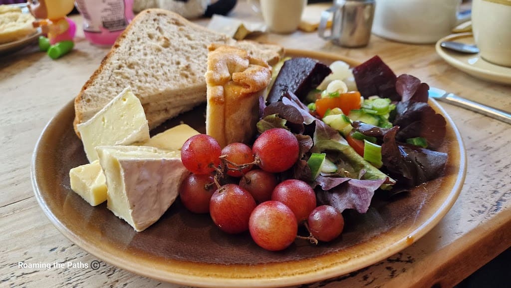 
This image shows a close-up of a beautifully arranged ploughman’s lunch served on a brown ceramic plate. The focus is on a variety of fresh, traditional elements. In the foreground, a small bunch of red grapes adds a pop of color, nestled next to a rich, creamy wedge of Brie cheese. A golden mini pork pie sits near the center of the plate, providing a savory touch. Behind the grapes and cheese, slices of rustic whole-grain bread are stacked neatly.

The right side of the plate features a colorful salad made of mixed greens, including dark purple lettuce, and garnished with sliced spring onions. Next to the salad, cubes of beetroot and pickled onions add a tangy flavor. The plate is set on a light wood table, with the edge of a knife peeking into the frame on the right. In the blurred background, there are cups and a teapot, suggesting a cozy café setting perfect for enjoying a comforting meal like this. The overall presentation is both rustic and inviting, highlighting the variety of textures and flavors in the dish.