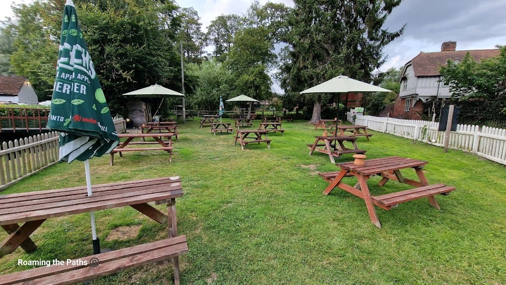 The image shows the spacious riverside garden of The Woolpack in Sopley, featuring a neatly kept grassy area with several wooden picnic benches scattered across the lawn. Some of the benches are shaded by large green umbrellas, providing a pleasant spot for outdoor dining or relaxing.

The garden is bordered by a white picket fence on one side, with large trees and greenery in the background, creating a peaceful, natural setting. The area appears open and inviting, perfect for enjoying the fresh air and surroundings. In the distance, there is a small wooden shed, and traditional English buildings can be seen beyond the fence, adding to the rustic charm of the setting.