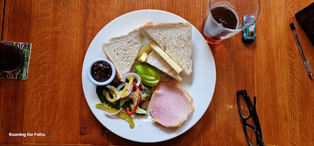 
This image shows a flat-lay view of a classic Ploughman’s lunch served on a white plate, resting on a wooden table. The meal includes several traditional components arranged neatly across the plate. In the upper portion, two slices of plain white sandwich bread are stacked on top of each other. Tucked under the bread, there are two slices of cheddar cheese and a slice of brie cheese, creating a creamy contrast to the sandwich-style bread.

To the left, a small white ramekin holds a generous portion of dark chutney, which looks rich and tangy, ready to complement the cheese and bread. Beside the chutney is a fresh, colorful salad with mixed greens, sliced red onions, and strips of red and yellow bell peppers, lightly drizzled with a mustard dressing. The salad adds a vibrant, fresh element to the otherwise rich and hearty plate.

In the lower right corner, there is a thick slice of ham with a slightly pink center and well-defined edges, which appears to be home-cooked or freshly carved, contributing to the traditional feel of the meal. Next to the ham, there are slices of crisp green apple, adding a sweet and tangy balance to the dish.

In the background, you can see a half-filled pint glass of what appears to be ale, placed to the top right of the plate. A pair of black eyeglasses and a pen sit to the right of the plate, suggesting a relaxed pub setting. The rich brown, wooden texture of the table adds a rustic touch, enhancing the hearty, comforting nature of this traditional British pub meal. Additionally, a colorful drink coaster with a partial glass of a dark-colored beverage is visible to the left, further completing the atmosphere of a cozy dining experience.