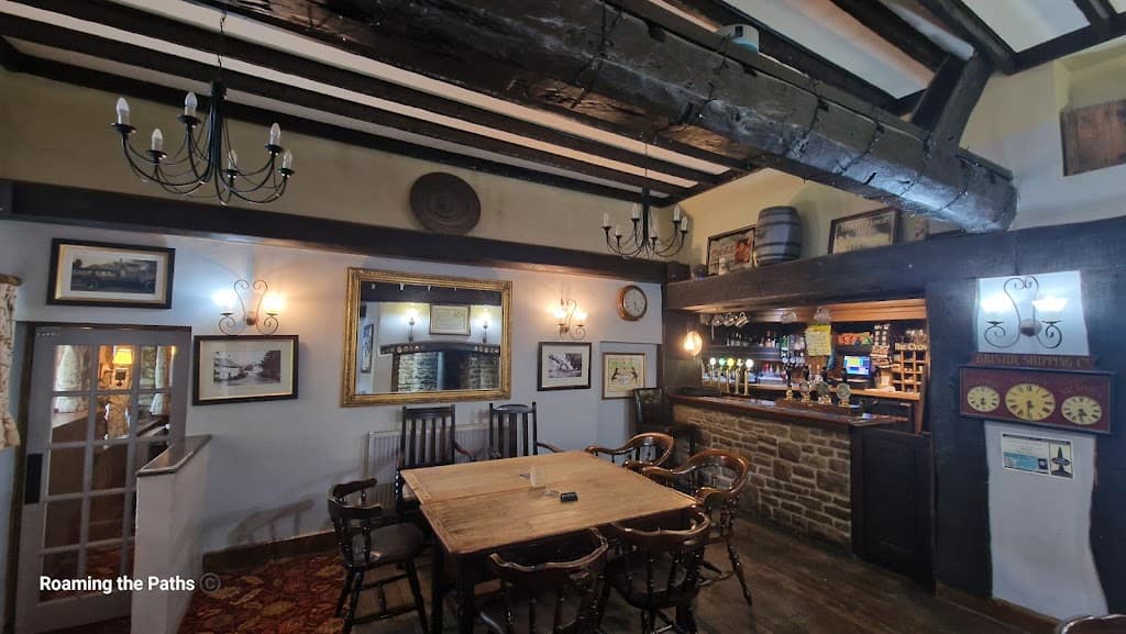 The image shows the cozy, rustic interior of the Crown Inn at Puncknowle, a traditional English country pub. The focal point is a large wooden table surrounded by dark wooden chairs with arms, providing an inviting spot for patrons to gather. Above, thick wooden ceiling beams span the room, adding a historic, rustic charm. Two black metal chandeliers with candle-style lights hang from the ceiling, further emphasizing the old-world ambiance.

In the background, the bar is neatly arranged with various beers on tap, highlighted by warm hanging lights. The bar front is made of stone, and a shelf behind it displays various bottles, glasses, and pub décor. On the wall to the left of the bar, vintage signs and framed paintings hang, while a large, golden-framed mirror reflects the room, adding depth. Above the bar, traditional pub signs and a decorative wooden barrel are displayed, giving the space a sense of heritage and authenticity.

A clock and framed artwork line the back walls, adding to the pub's homey feel. The carpet has a classic design, complementing the warm tones of the wood and stone. The overall atmosphere is inviting and reminiscent of a traditional English pub, with a sense of history and a welcoming vibe for visitors and locals alike.