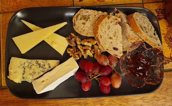 This flatlay image showcases a neatly arranged cheese platter on a black rectangular plate. The cheeses include a sharp cheddar cut into triangular wedges, a creamy wedge of brie, and a piece of blue cheese with strong veining. Surrounding the cheese are several slices of artisan bread, likely containing olives or fruit pieces, adding texture and color. A handful of walnuts, a small cluster of red grapes, and a glass bowl filled with dark chutney complete the platter. The table beneath has a rustic, wooden finish, contributing to the cozy ambiance.