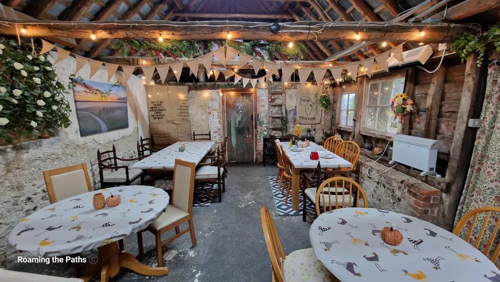 This image captures the rustic and cozy interior of The Old Forge farm shop in Berwick St James. The room has a warm, inviting ambiance with its exposed wooden beams, stone walls, and eclectic decor. The tables are covered in a whimsical tablecloth featuring colorful chicken illustrations, adding a touch of country charm.

String lights and triangular bunting are draped across the ceiling, creating a festive and homely atmosphere. Various tables are arranged with a mix of wooden and cushioned chairs, providing a relaxed seating arrangement. The walls are adorned with burlap coffee sacks, vintage posters, and decorative plants, enhancing the rustic theme. A large, faded wooden door adds character to the space, while a atumnal wreath  by the window adds a pop of color. This setting is perfect for enjoying a leisurely meal or tea, surrounded by rustic charm and a cozy atmosphere.