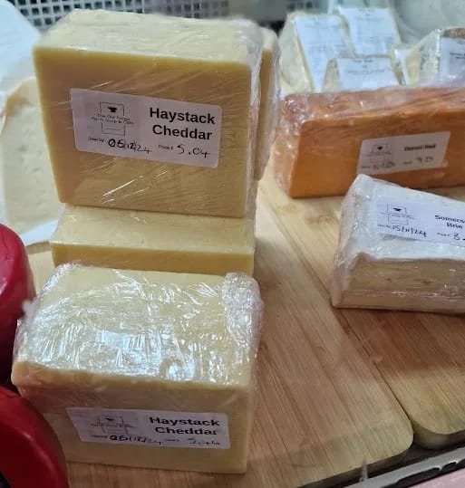 This image displays a selection of cheese blocks at a farm shop or deli, specifically focusing on Haystack Cheddar. The blocks of cheddar are neatly wrapped in plastic, each with a label indicating the name "Haystack Cheddar" along with weight, price, and sell-by date.

The cheddar is a pale yellow color, characteristic of classic cheddar, with a firm, smooth texture visible through the wrapping. The display also includes other varieties of cheese in the background, some with a deeper yellow or orange hue, suggesting different types or aging levels. The cheeses are arranged on a wooden board, adding to the rustic and artisanal feel of the presentation.