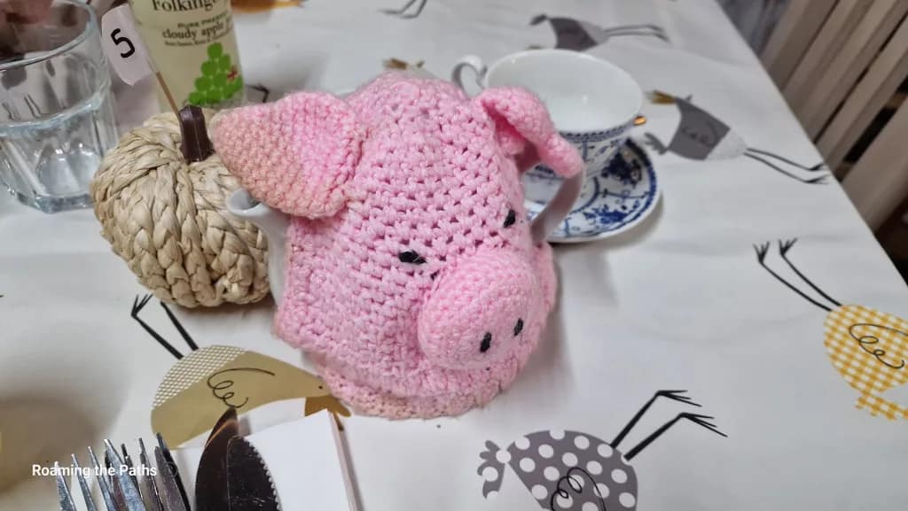 This image showcases a charming pink crocheted tea cosy in the shape of a pig, complete with a snout, floppy ears, and embroidered eyes. The tea cosy covers a teapot, creating a whimsical and cozy touch to the table setting at The Old Forge farm shop.

The cosy sits on a farm-themed tablecloth decorated with illustrations of chickens in various colors, adding to the rustic, country ambiance. In the background, there are items like a wicker napkin holder, a cup and saucer with a blue and white pattern, and a bottle of Folkington’s cloudy apple juice. This delightful setup combines warmth and character, perfect for a cozy meal or afternoon tea at the farm shop.