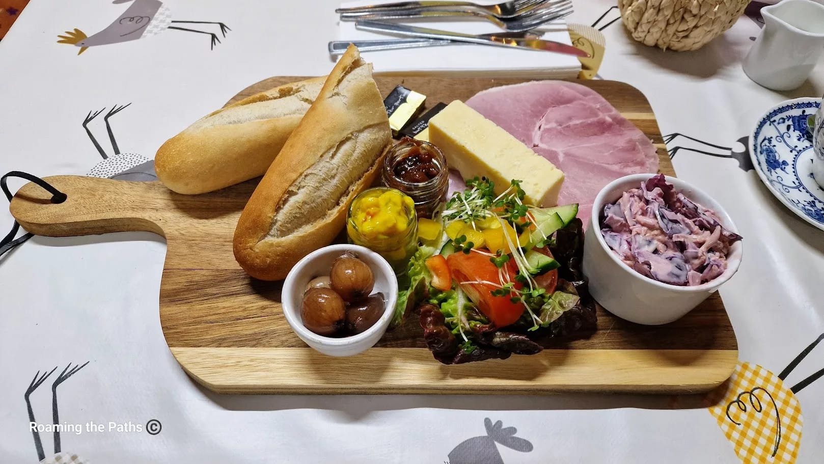 This image shows a traditional Ploughman's Lunch served on a wooden board at The Old Forge farm shop. The presentation includes a variety of elements: Two small baguette halves – Fresh, crusty baguette segments, ready to be paired with other items on the board. Thick slice of cheddar cheese – A wedge of creamy, yellow cheddar, a staple in classic Ploughman’s fare. Sliced ham – Large, thin slices of ham, with a slightly pink hue and tender texture, lying next to the cheese. Condiments and accompaniments: A small jar of chutney (possibly caramelized onion or fruit-based). A small jar of piccalilli – a bright yellow, tangy pickle relish with chunks of vegetables. A small bowl of pickled onions – glazed and shiny, adding a sour punch to the meal. Mixed salad – A fresh side salad featuring lettuce, tomatoes, cucumber, and sprouts, arranged neatly in one corner. Creamy coleslaw – A small bowl of coleslaw with a pinkish tint, likely from the inclusion of red cabbage, adding a crunchy and creamy element to the plate. The board is set on a farm-themed tablecloth, with whimsical chicken illustrations, and cutlery is arranged in the background. The entire presentation has a rustic and hearty feel, fitting for a classic British farm shop meal.
