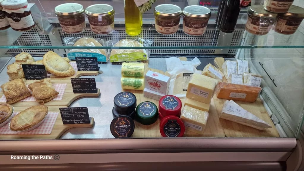 This image shows a well-stocked deli counter featuring an assortment of locally sourced pies, cheeses, and gourmet spreads.

Pastries and Pies – On the left side, there are various savory pastries, including pork pies, chicken and bacon pasties, and other homemade treats. Each item is labeled with ingredients, allergens, and pricing for both dine-in and takeaway options.

Cheeses – The counter displays a variety of cheeses, including blocks of Haystack Cheddar, Dorset Red (noted for its smoky flavor), and Snowdonia Cheese in black and red wax rounds, offering a selection of flavors for cheese enthusiasts.

Butter and Spreads – In the center, there are packs of salted and unsalted farmhouse butter, adding to the artisanal selection. Along the top shelf are jars of Gourmico pâtés and chutneys, perfect accompaniments for the cheeses.

The display is neatly organized on wooden boards, giving a rustic feel. The range of local and specialty products invites customers to enjoy the rich, savory flavors of this traditional deli.