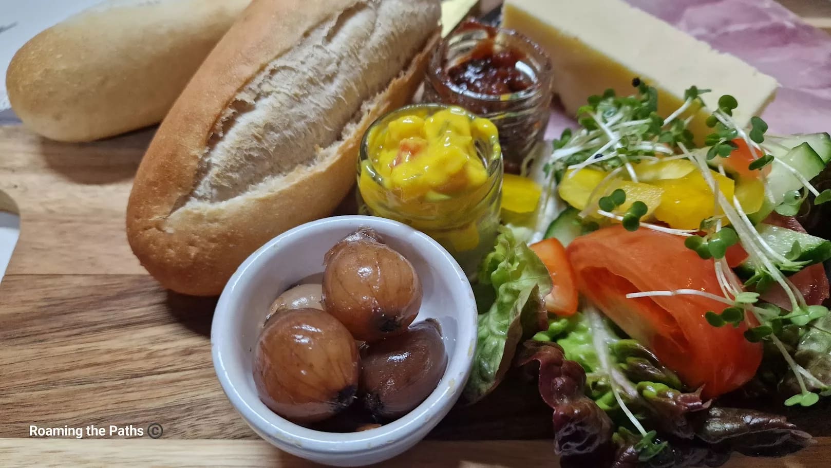 This image is a close-up of a portion of the Ploughman’s Lunch, emphasizing the fresh accompaniments, condiments, and bread. Here are the detailed components shown:

Bread – A crusty, fresh half-baguette, sliced open, perfect for pairing with cheese, ham, or condiments.
Pickled onions – Small, glossy, pickled onions served in a white bowl, offering a sharp, tangy flavor.
Piccalilli – A small jar filled with bright yellow piccalilli, a classic relish made of chunky pickled vegetables in a mustard sauce, adding a tangy and slightly spicy contrast.
Chutney – A small jar of chutney, possibly onion or fruit-based, providing a sweet and savory accompaniment.
Side salad – A fresh salad featuring slices of tomato, cucumber, yellow bell pepper, lettuce, and microgreens, adding a crisp, vibrant element to the dish.
The close-up captures the textures and colors vividly, showcasing the contrast between the rich condiments, fresh vegetables, and hearty bread, highlighting the essence of a traditional and wholesome Ploughman's Lunch.